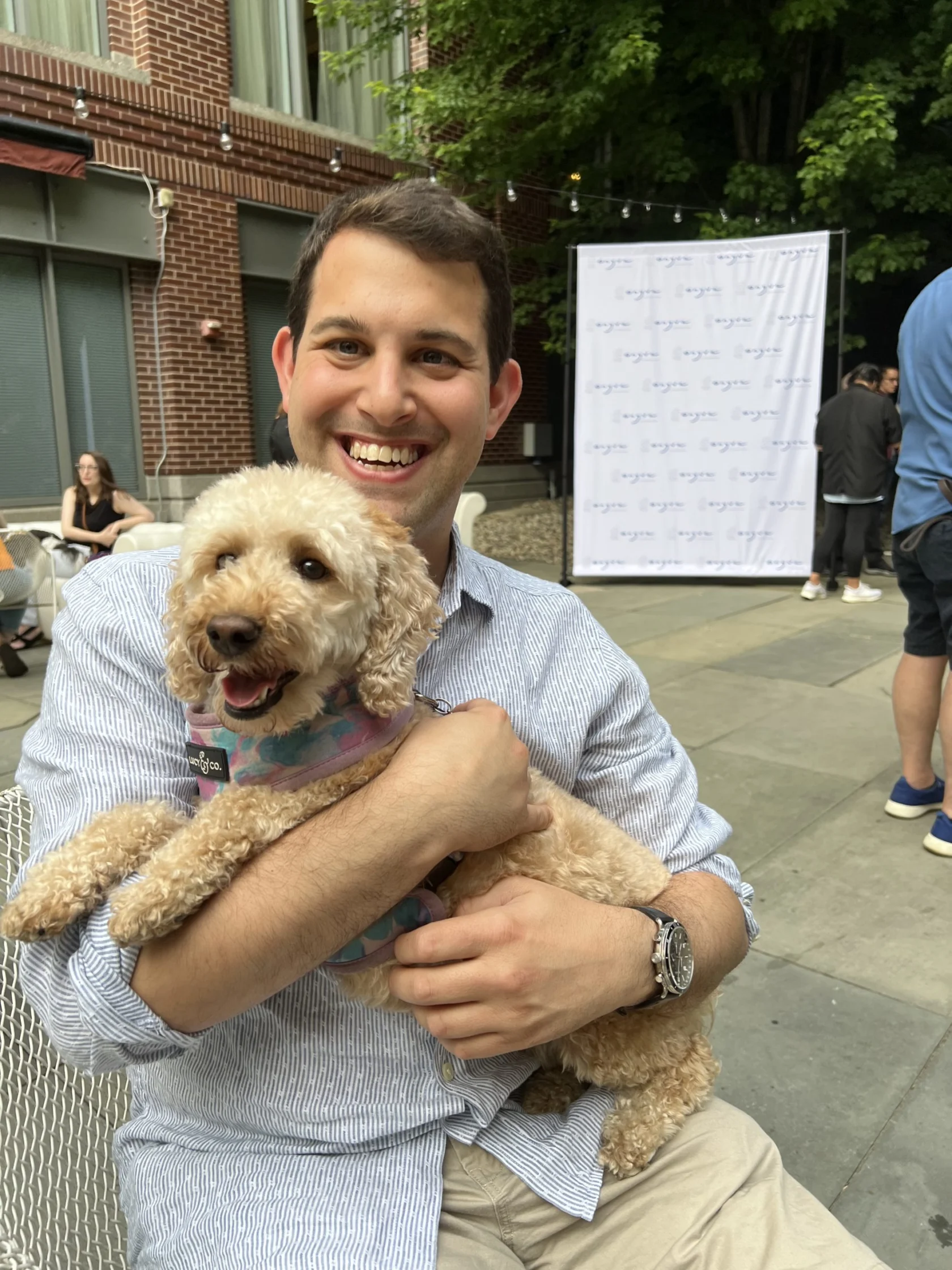 A man sitting outdoors smiles while holding a small curly-haired dog in his lap. Other people are in the background near a white backdrop.