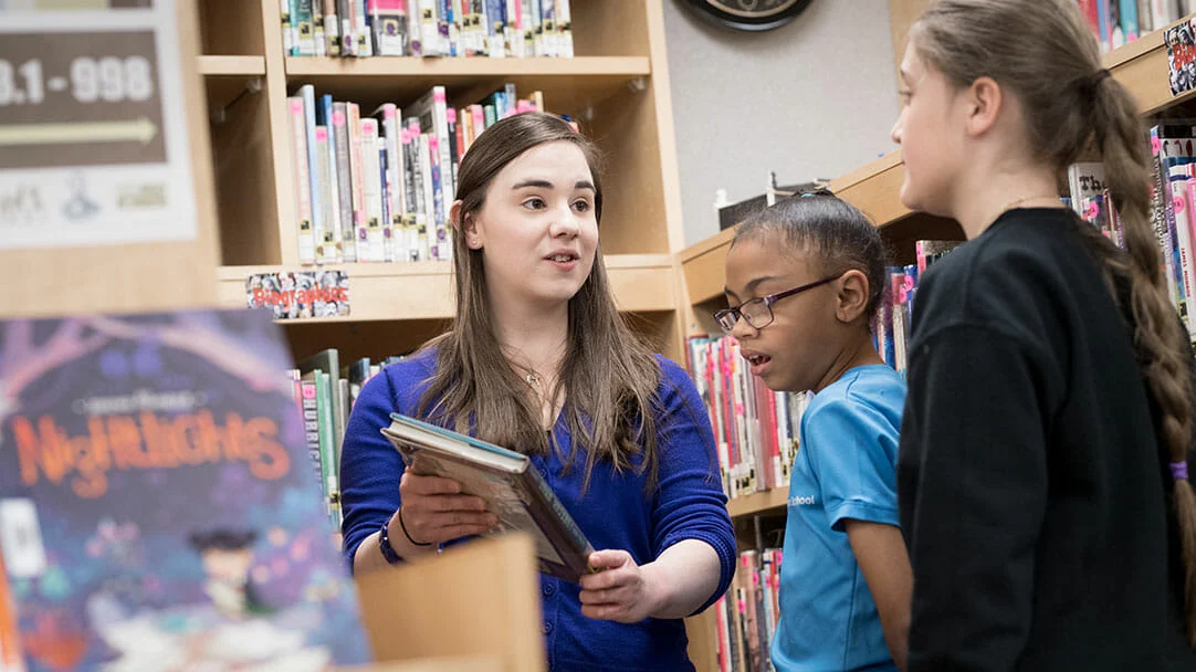 A woman holding books speaks to two children in a library, surrounded by bookshelves and books.