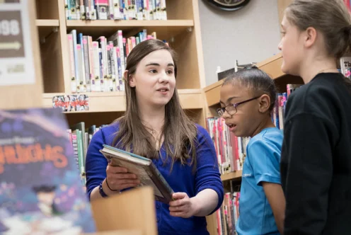 A woman holding books speaks to two children in a library, surrounded by bookshelves and books.