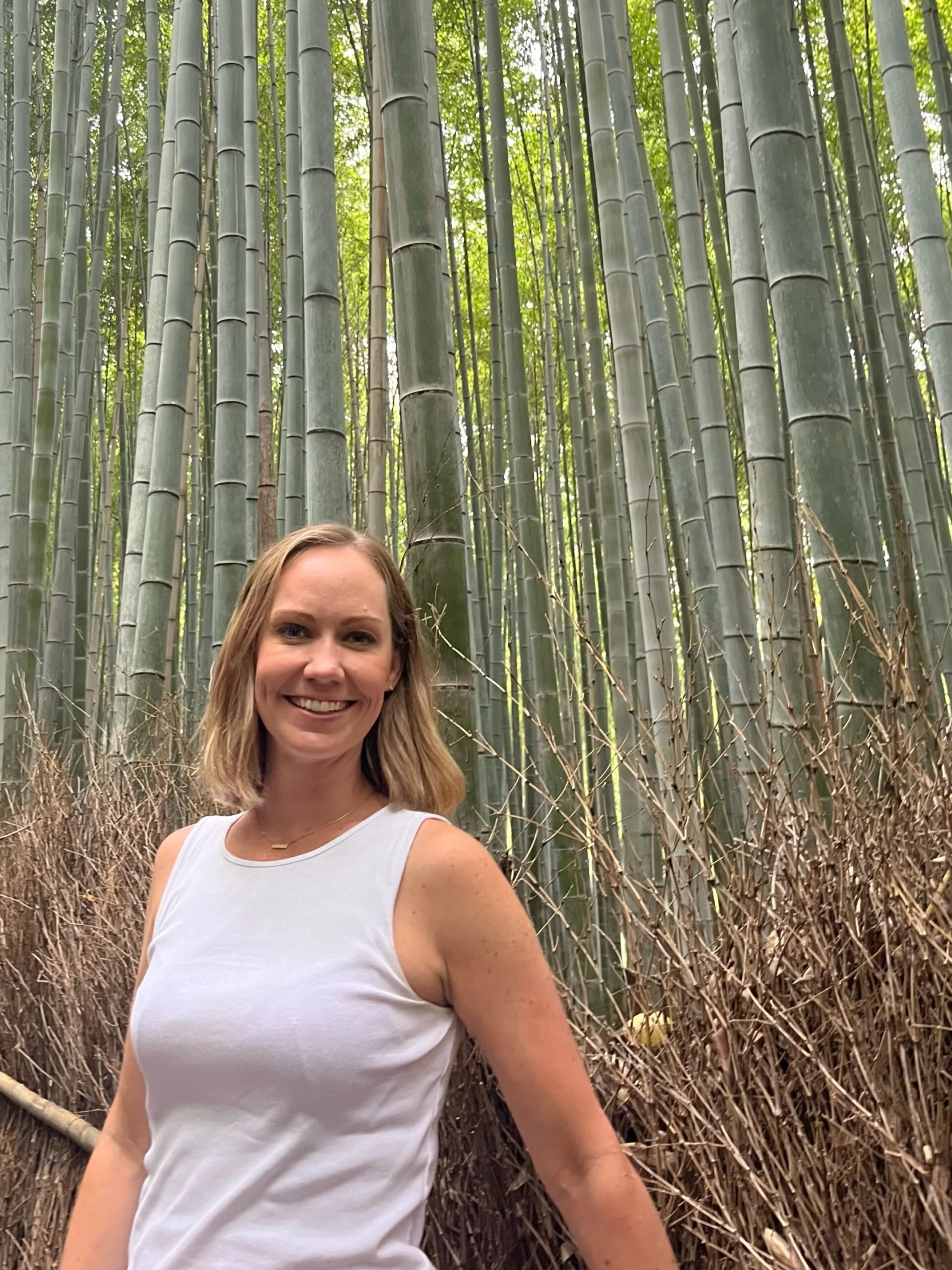 A woman in a white sleeveless top stands smiling in front of tall bamboo stalks in a dense bamboo forest.