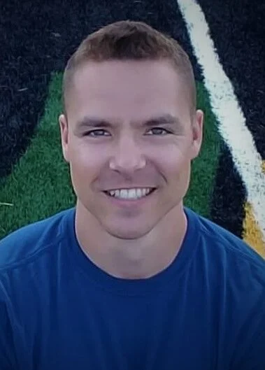 A man with short brown hair and a blue shirt smiles at the camera while sitting on a sports field with black, green, and yellow lines.