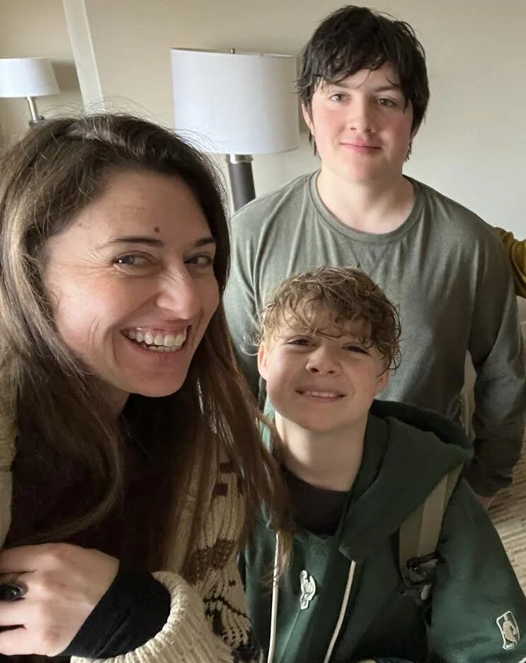 Three people are indoors smiling at the camera; an adult and two children stand close together in a room with beige walls and a lamp in the background.
