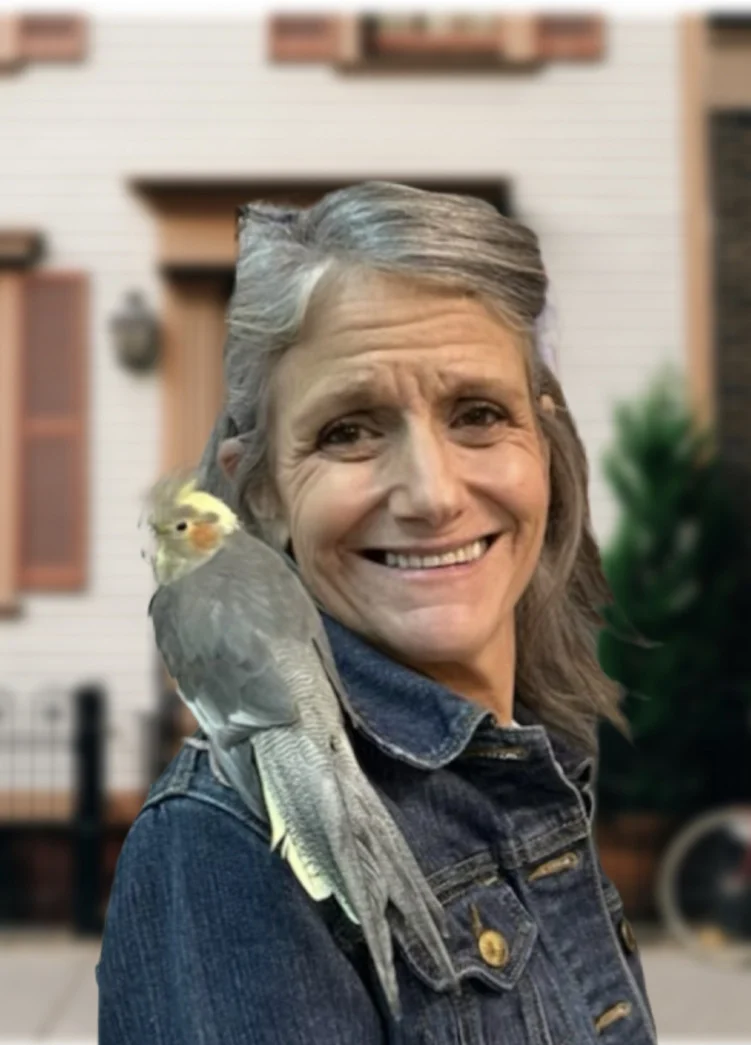 A woman with gray hair wearing a denim jacket smiles at the camera with a cockatiel perched on her shoulder, standing in front of a house.