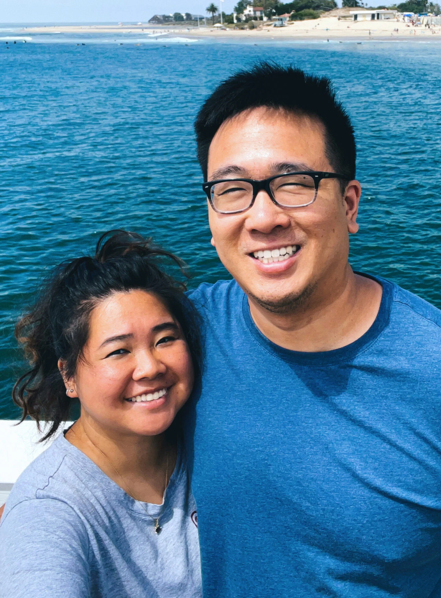 Two people stand close together smiling in front of a body of water, with a distant shoreline, beach, and trees visible in the background.