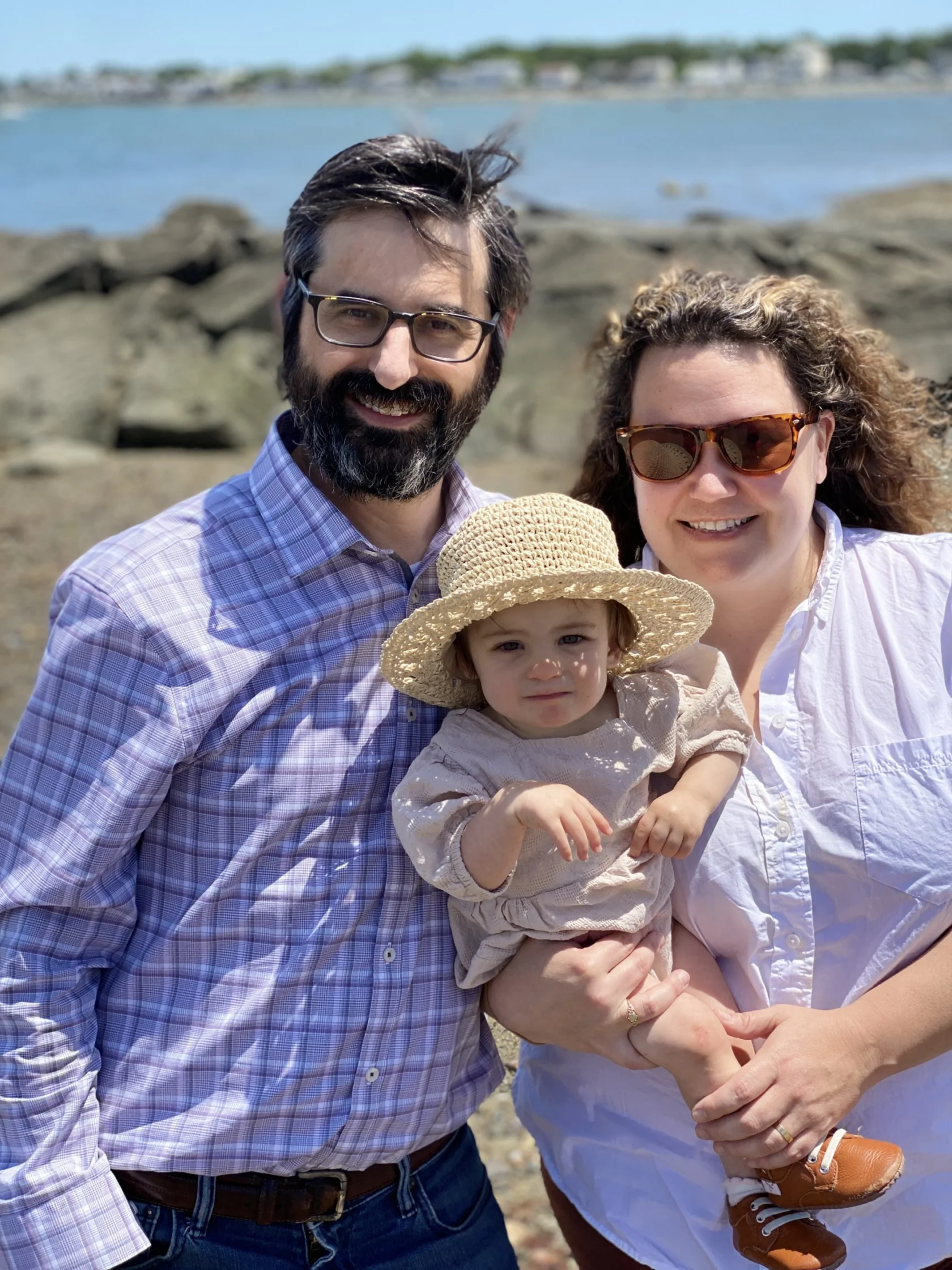 A man, woman, and young child stand together on a rocky shoreline with water and distant buildings in the background. The child wears a straw hat.