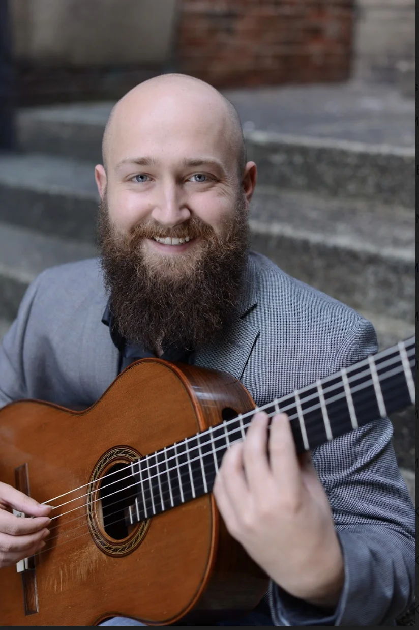 A bearded man in a gray suit sits on outdoor steps, smiling while playing a classical acoustic guitar.