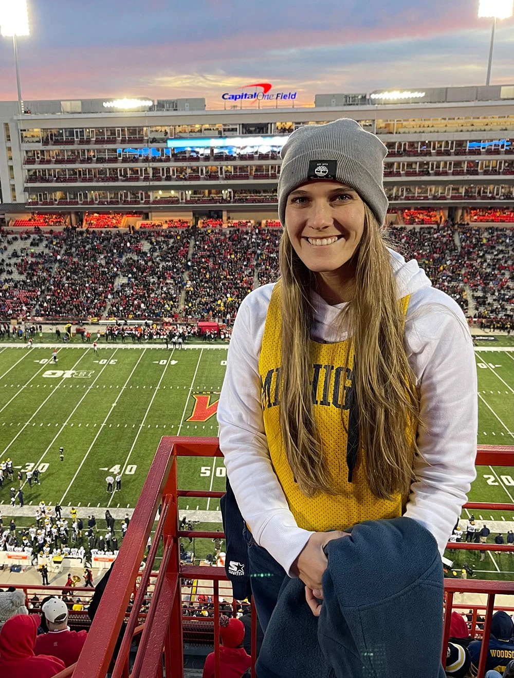 A woman wearing a gray beanie and a Michigan vest stands in a stadium during a football game, with the field and crowd visible in the background.