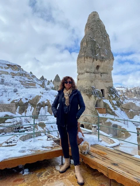 Person in winter clothing stands on a wooden platform with snow-covered rock formations and cloudy sky in the background, likely in Cappadocia, Turkey.