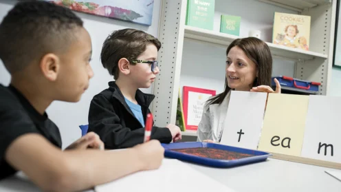 A teacher shows letter cards to two young students at a table, helping them learn to read in a classroom setting.