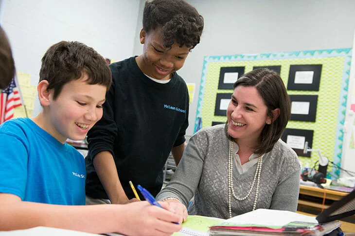 A teacher sits at a desk with two students, helping them with their work. One student writes while the other and the teacher watch and smile.