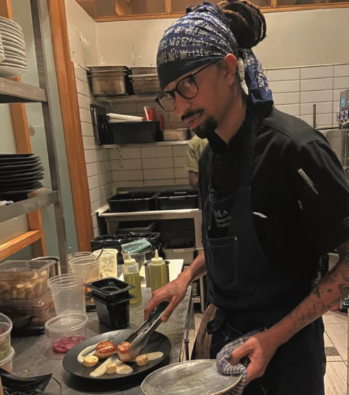 A chef wearing glasses and a blue bandana cooks food on a stove in a commercial kitchen, holding tongs and a plate.