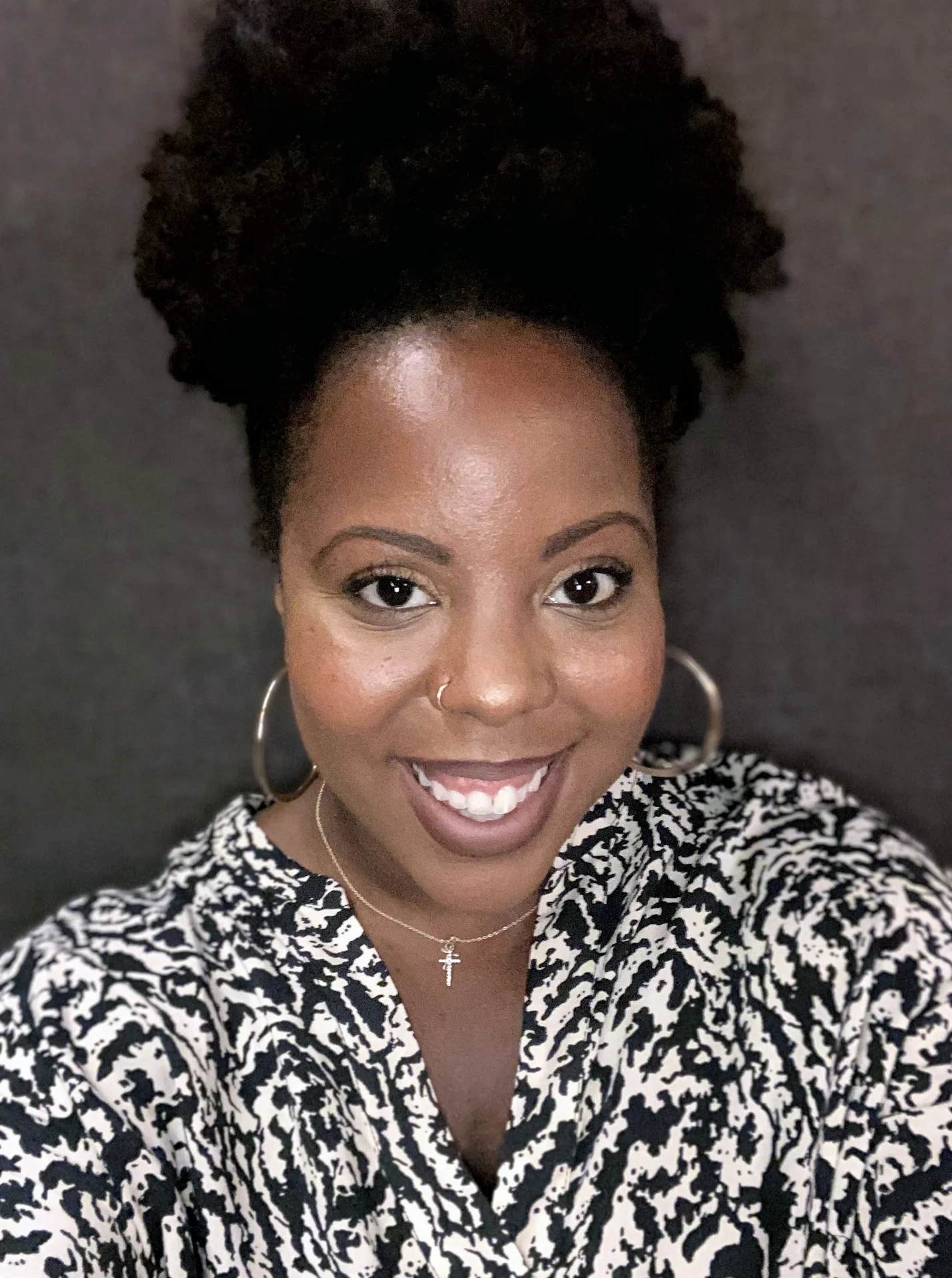 A woman with curly hair pulled up, wearing hoop earrings, a nose ring, a cross necklace, and a patterned top, smiles at the camera against a neutral background.
