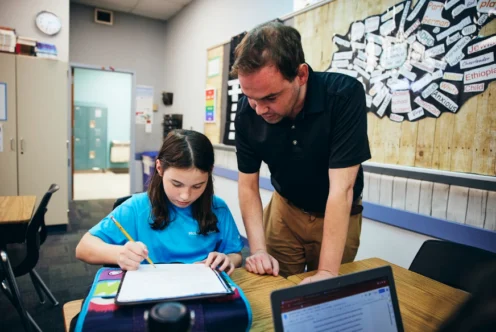 A teacher stands beside a seated student who is writing in a notebook, offering guidance in a classroom with educational materials on the wall.