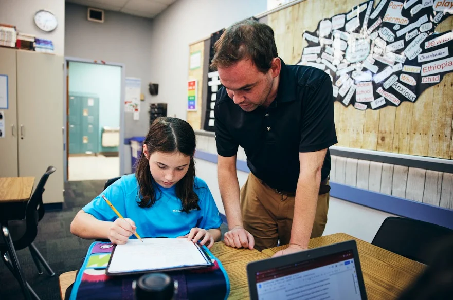 A teacher stands beside a seated student who is writing in a notebook, offering guidance in a classroom with educational materials on the wall.
