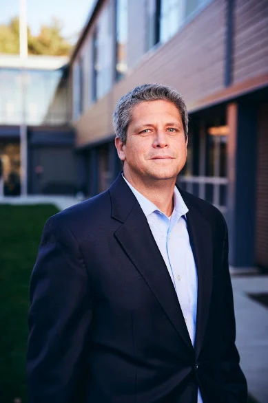 A man in a dark suit jacket and light blue shirt stands outside in front of a modern building, looking at the camera with a neutral expression.
