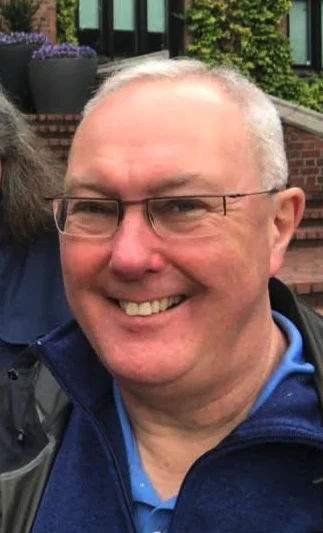A middle-aged man with short gray hair and glasses, smiling at the camera, wearing a blue shirt and jacket, standing outdoors in front of brick steps and green plants.