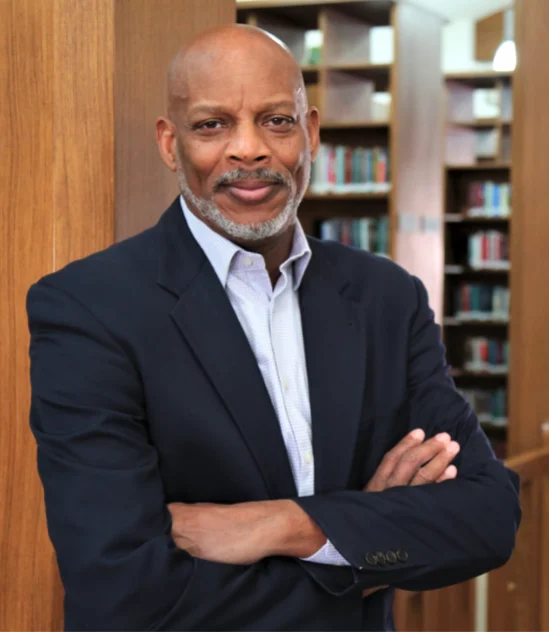 A man with a gray beard, wearing a dark suit and light shirt, stands with arms crossed in front of bookshelves in a library.