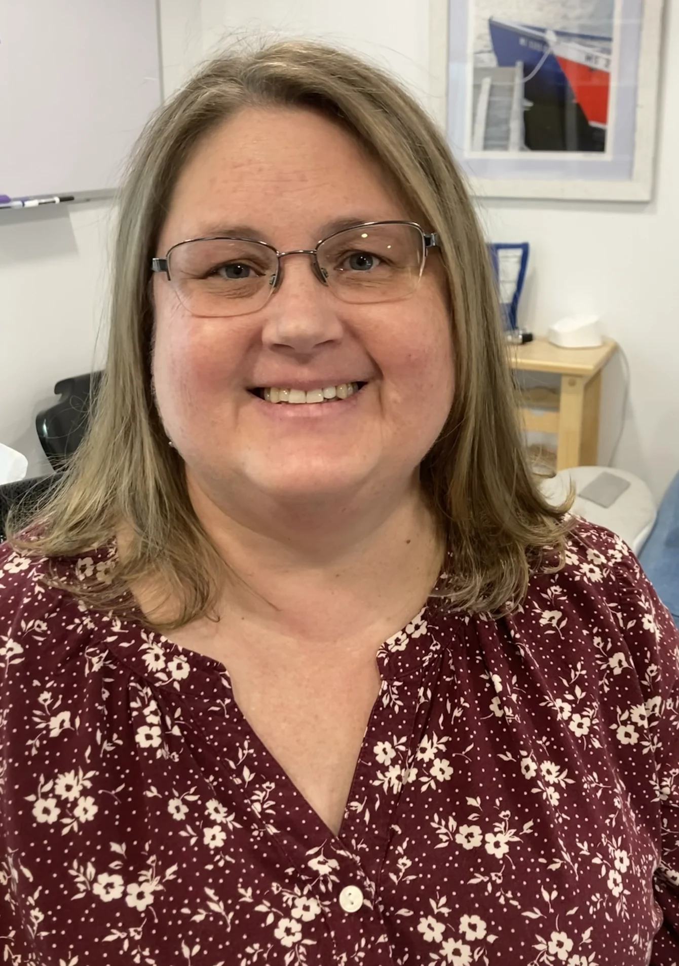 A woman with shoulder-length blonde hair and glasses smiles, wearing a maroon floral top, in an indoor office setting with a framed picture and whiteboard in the background.