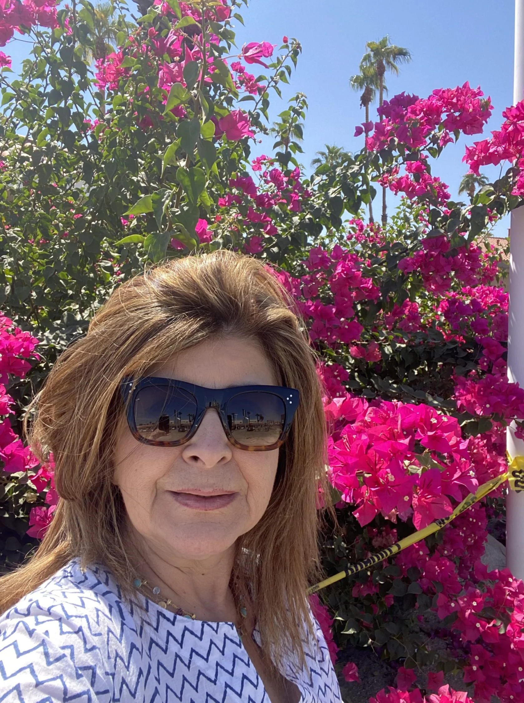 A woman wearing sunglasses and a patterned top stands in front of bright pink bougainvillea flowers on a sunny day.