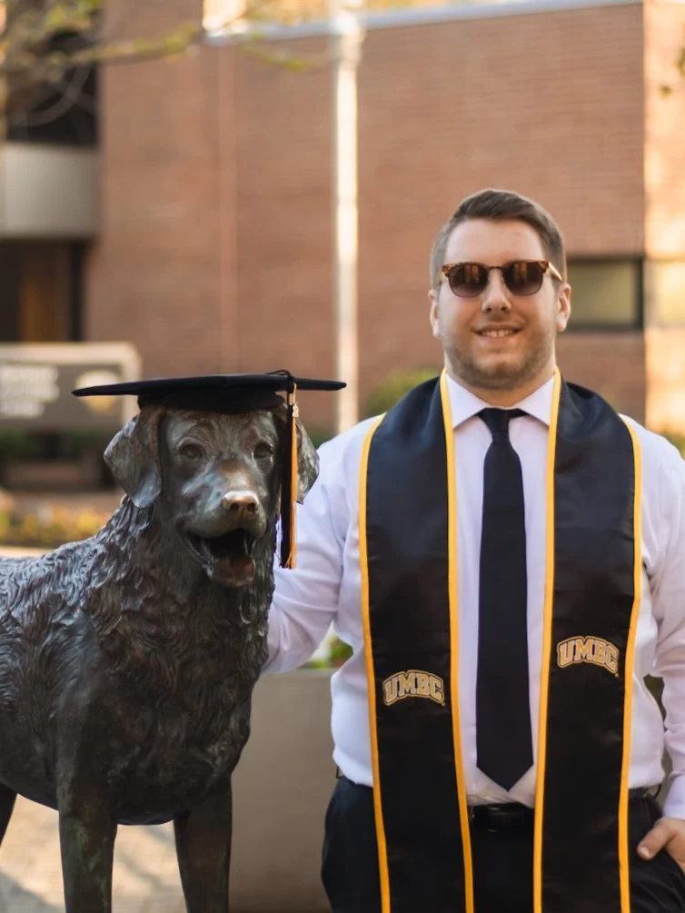 A man wearing sunglasses, a graduation stole, and tie stands next to a statue of a dog wearing a graduation cap, outdoors on a university campus.