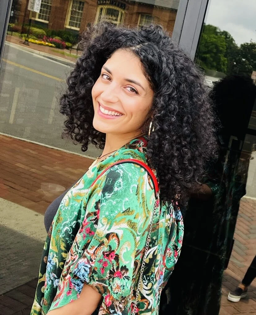 Woman with curly hair wearing a green floral-patterned kimono smiles at the camera while standing outside near a glass door.