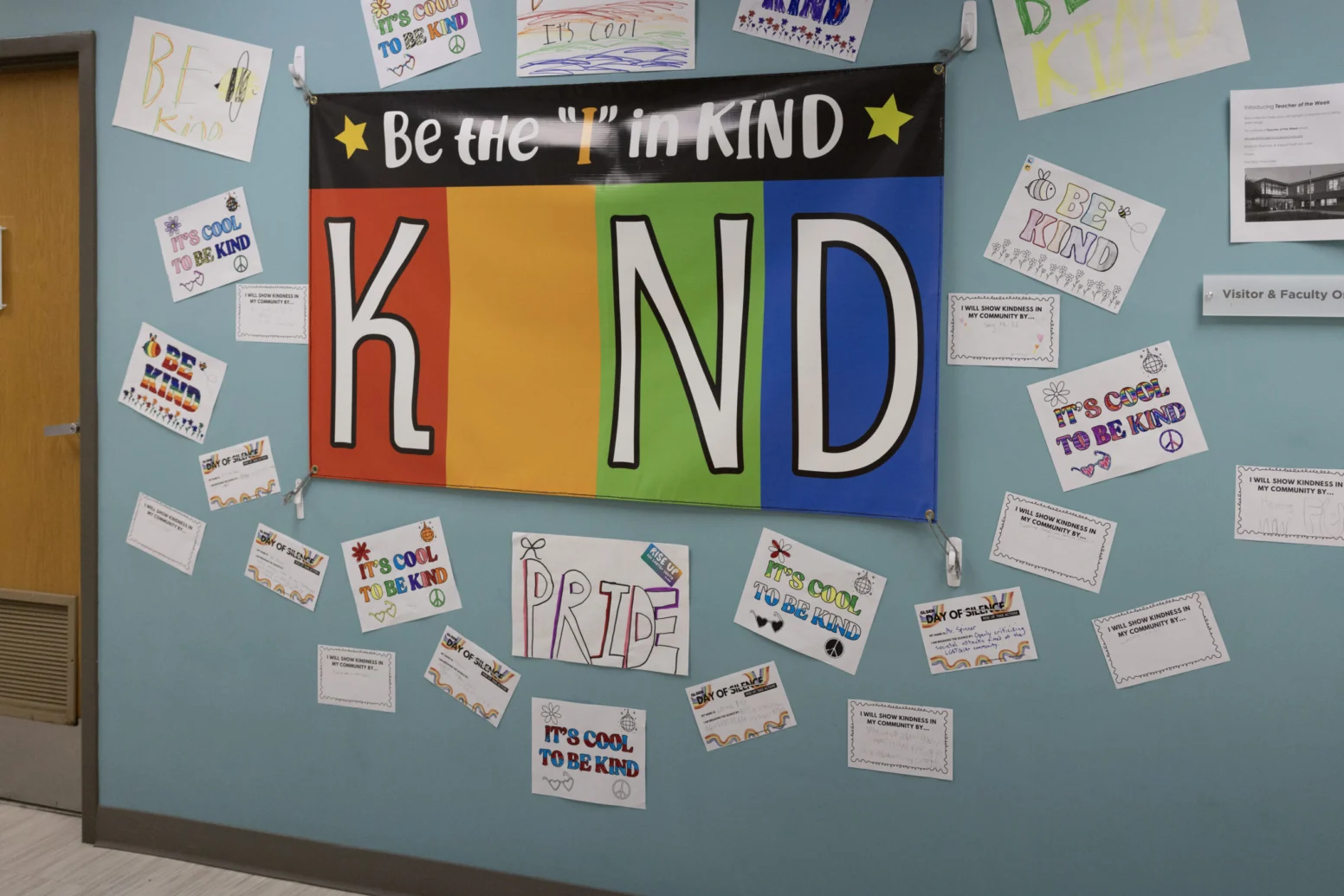 A colorful bulletin board displays a "Be the 'I' in KIND" banner surrounded by hand-drawn posters promoting kindness and pride.