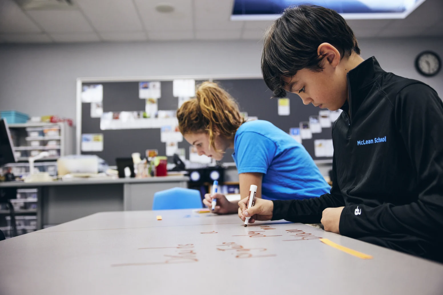 Two students write on their desks with markers in a classroom, focusing on their work. A bulletin board and shelves are visible in the background.