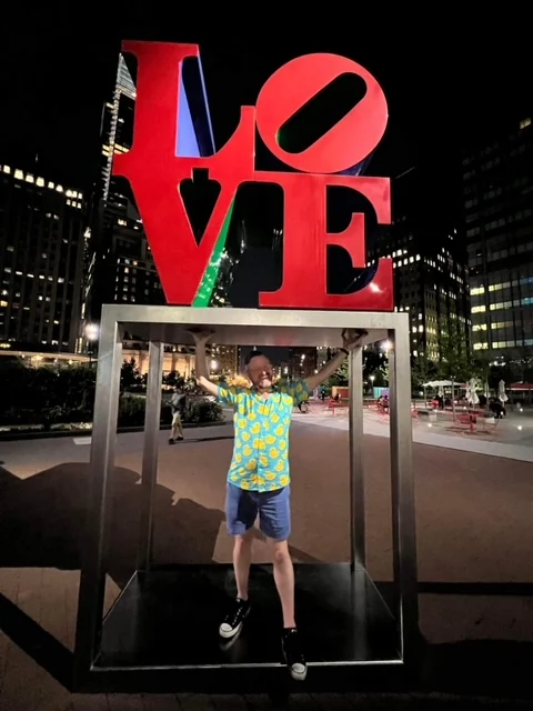 A person stands inside the frame of the LOVE sculpture at night, holding onto the bottom of the red letters, with buildings illuminated in the background.