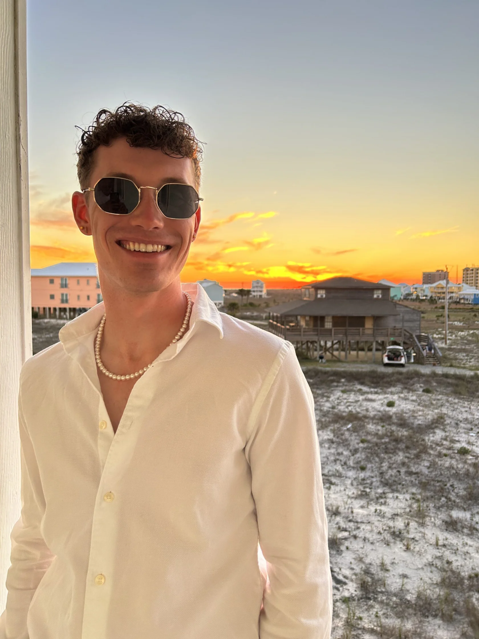 A man wearing sunglasses, a white shirt, and a pearl necklace stands outdoors, smiling, with a beach house and sunset in the background.