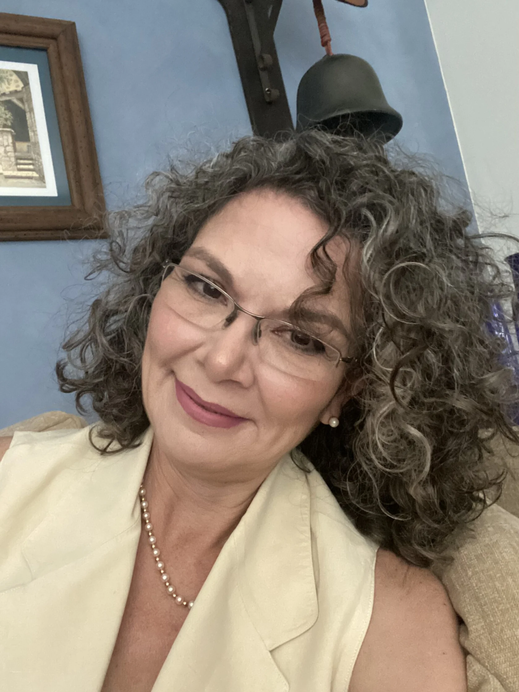A woman with curly gray hair, glasses, and a beige vest smiles at the camera. She wears a pearl necklace and sits indoors against a blue wall.