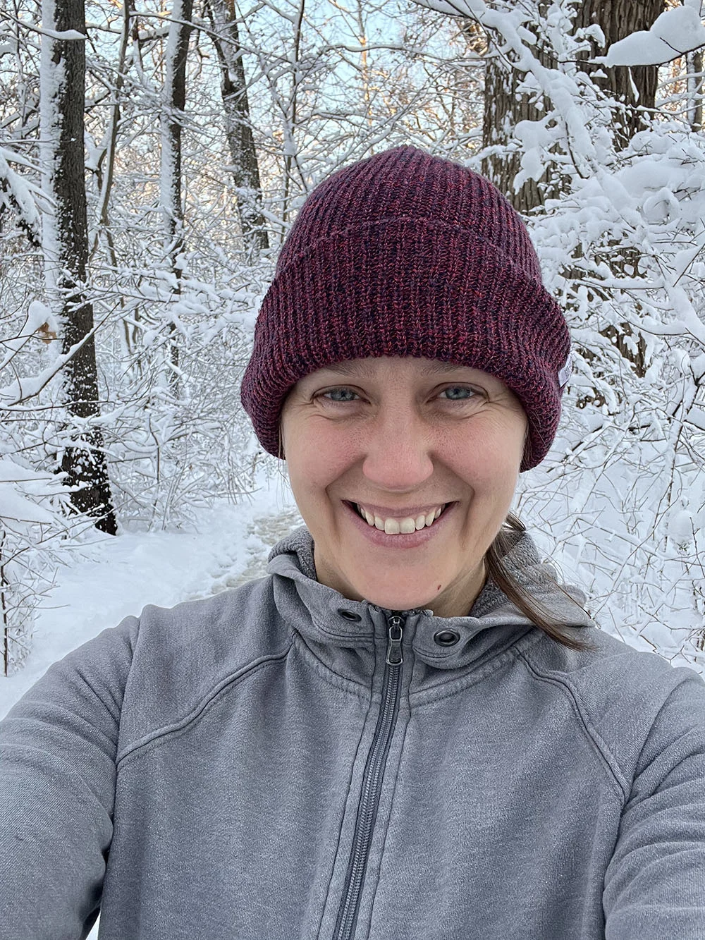 A person in a gray jacket and maroon knit hat smiles for a selfie on a snowy forest trail surrounded by snow-covered trees.