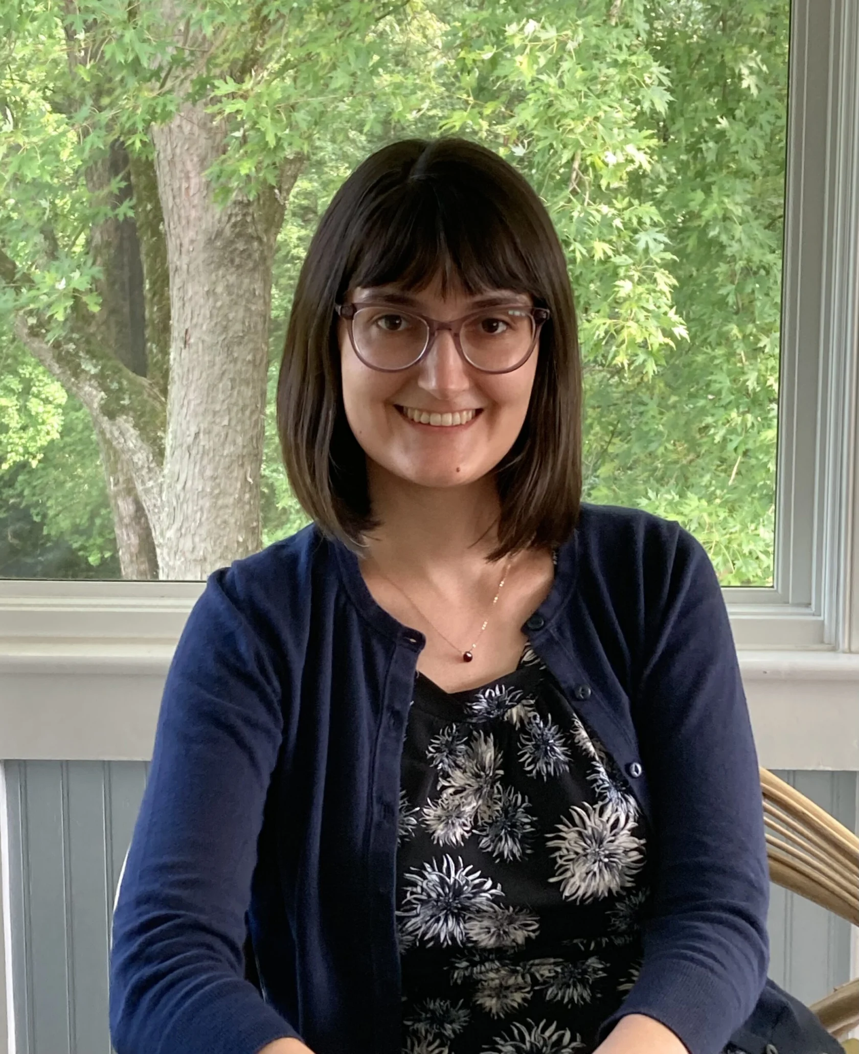 A woman with straight dark hair, glasses, and a navy sweater sits smiling in front of a window with green trees visible outside.
