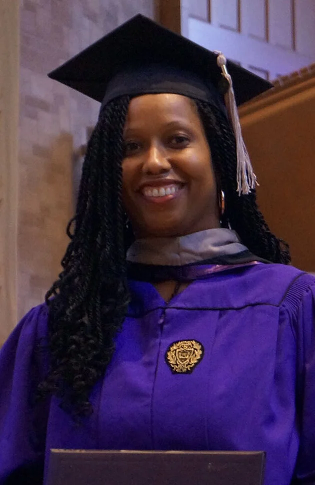 A woman wearing a graduation cap and gown smiles at the camera while holding a diploma inside a building.