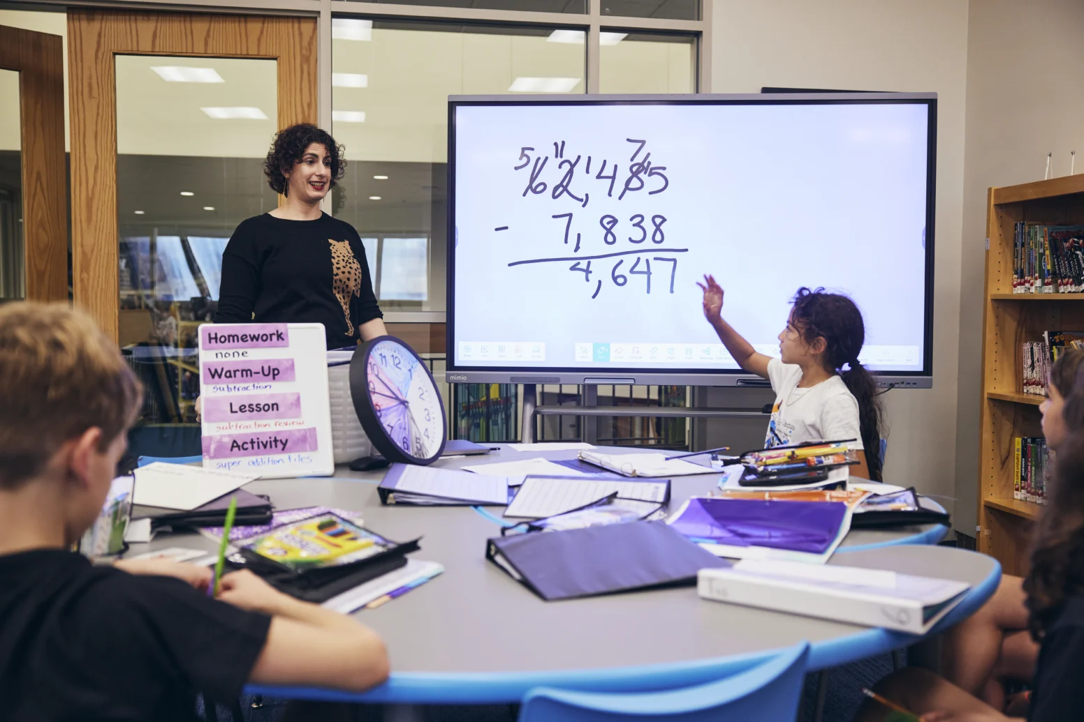 A teacher and students work on a math problem at a round table in a classroom, with a whiteboard displaying a subtraction equation in the background.