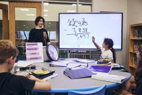 A teacher and students work on a math problem at a round table in a classroom, with a whiteboard displaying a subtraction equation in the background.