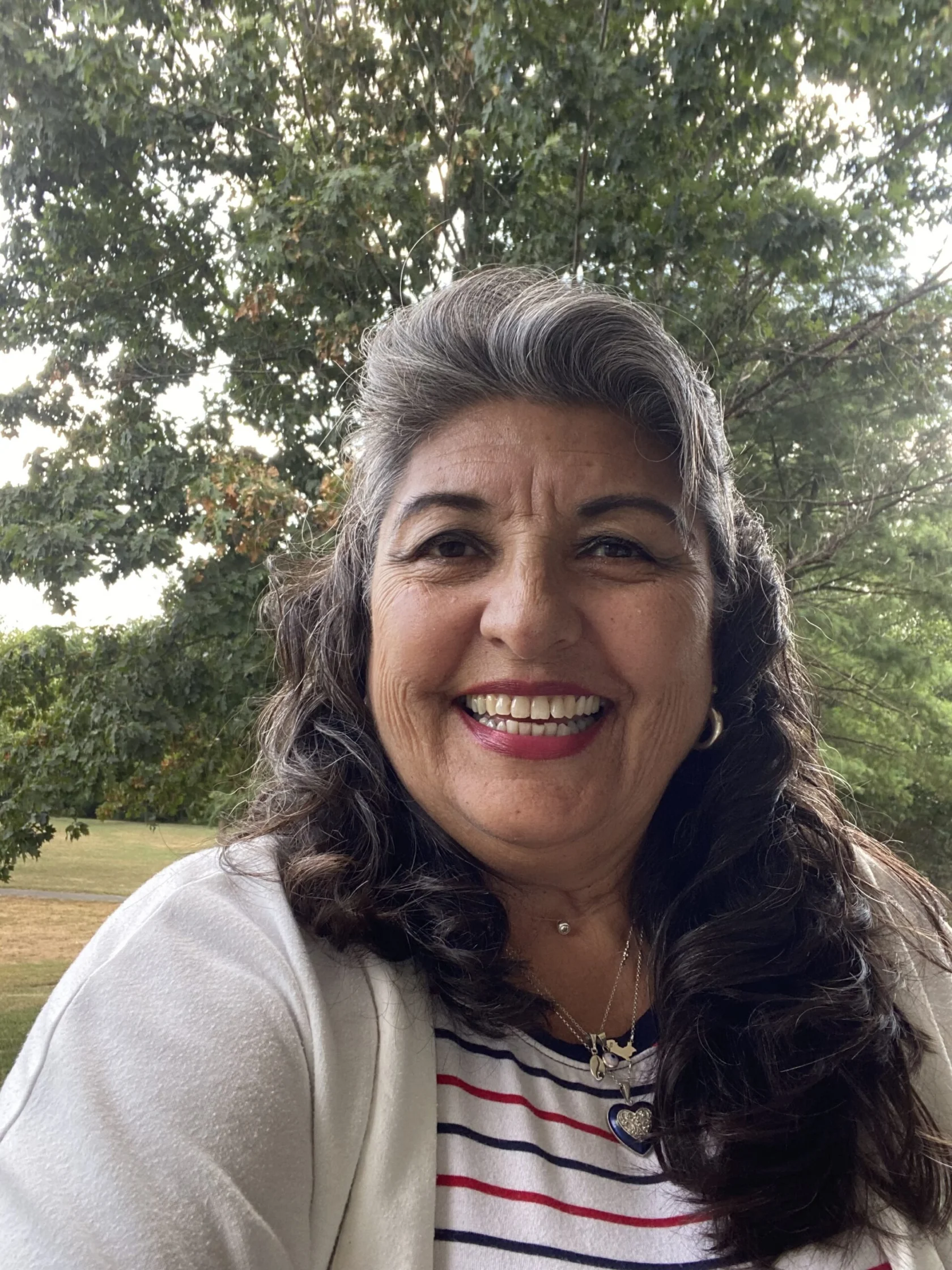 A woman with long curly hair smiles outdoors in front of green trees, wearing a white cardigan and a striped shirt.