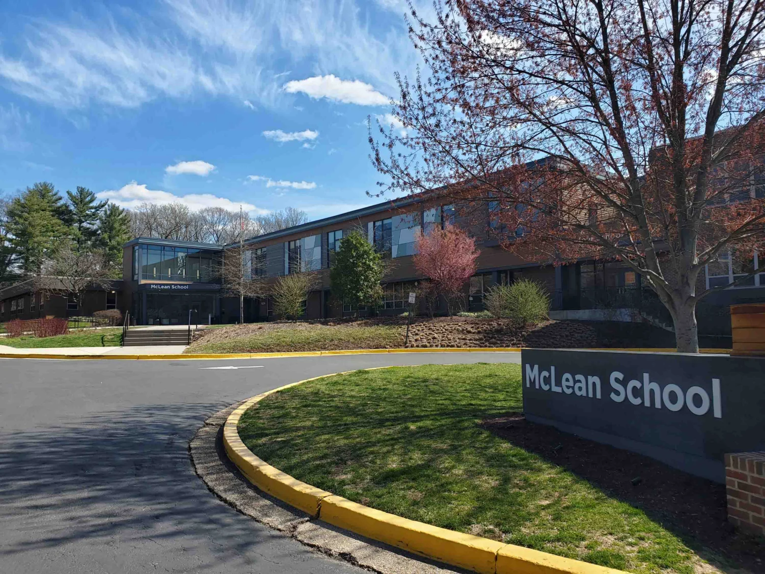 McLean School building with large windows, a sign in front, trees with spring foliage, and a blue sky with clouds.