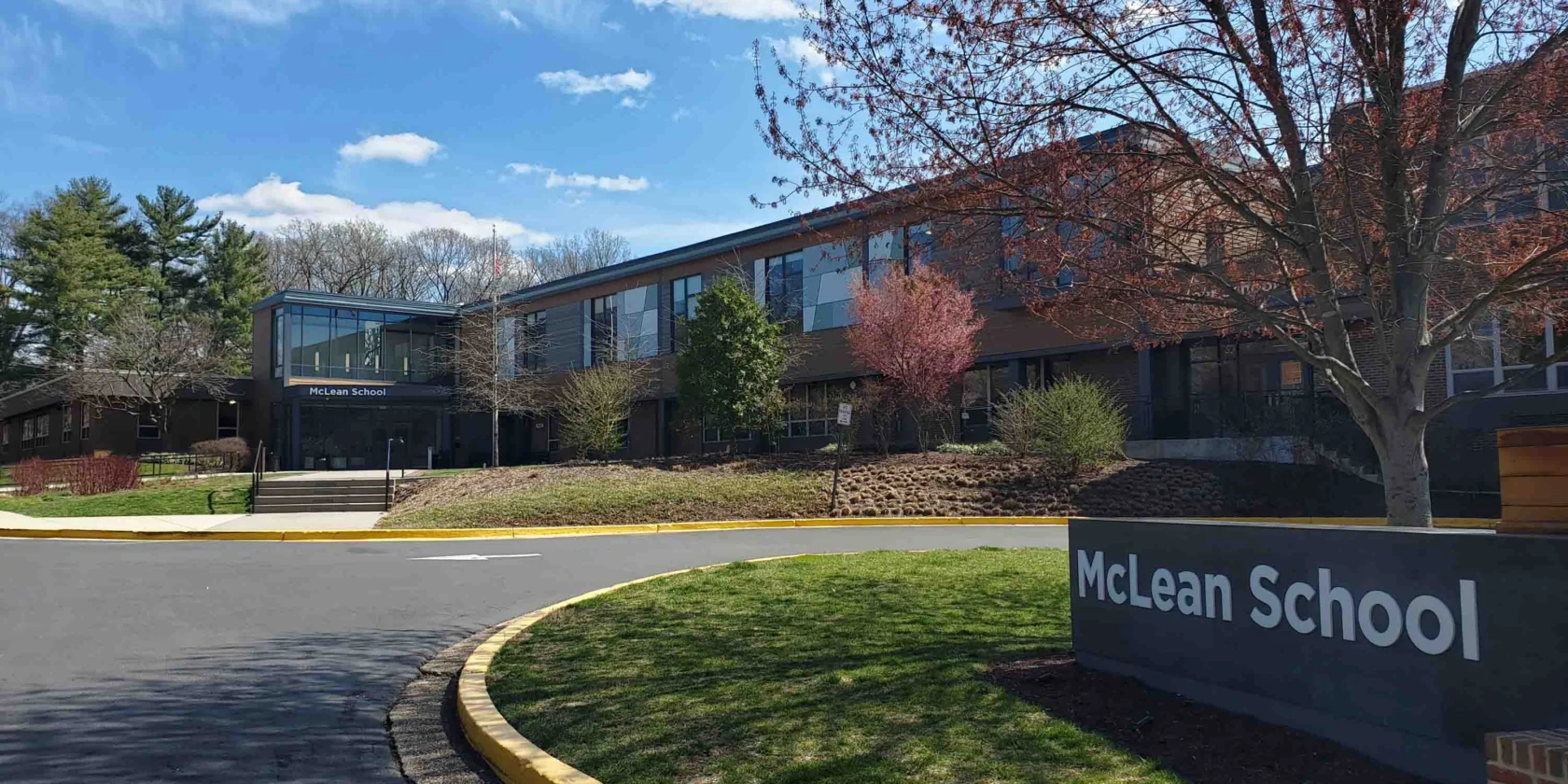 McLean School building with large windows, a sign in front, trees with spring foliage, and a blue sky with clouds.