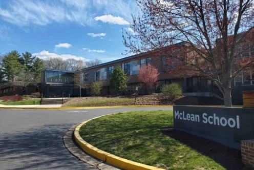 McLean School building with large windows, a sign in front, trees with spring foliage, and a blue sky with clouds.