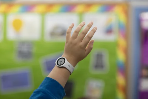 A child’s hand wearing a white wristband is raised in a classroom with a green bulletin board and colorful border in the background.