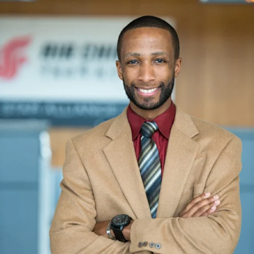 A man in a tan blazer, red shirt, and striped tie stands indoors with arms crossed, smiling at the camera. A blurred sign is visible in the background.