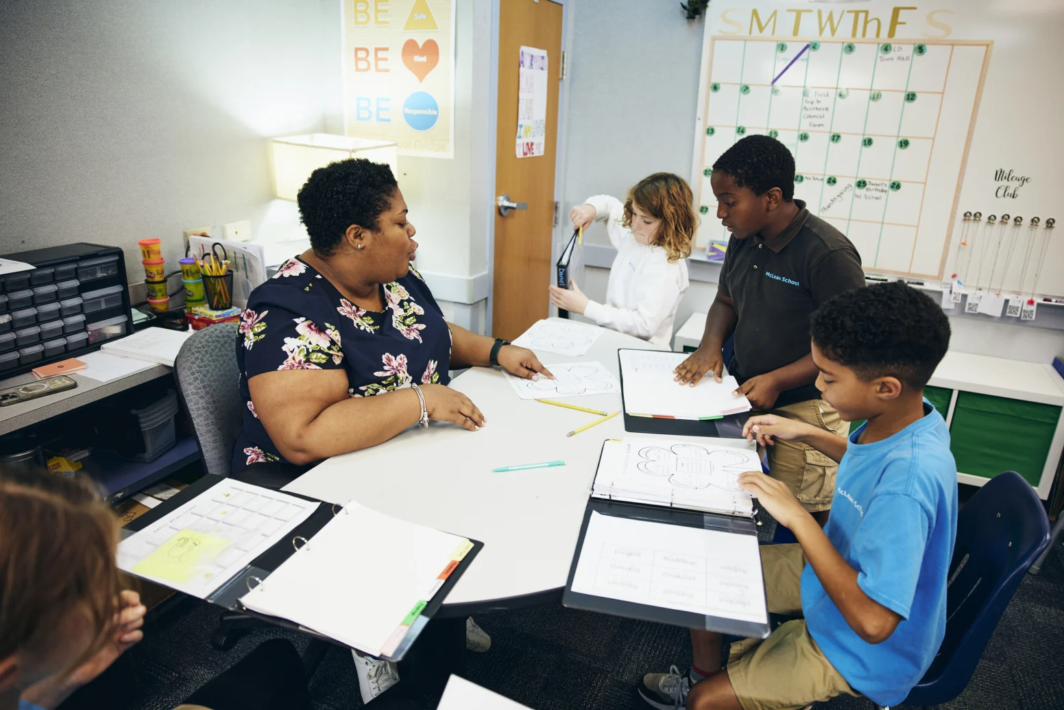 A teacher sits at a round table with four students, discussing worksheets and assignments in a classroom setting.