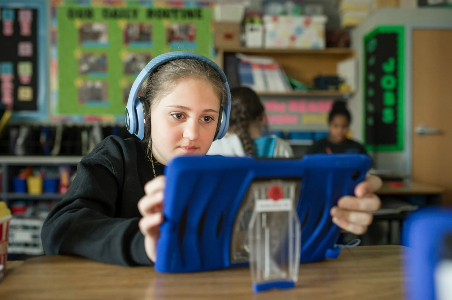 A student with headphones uses a tablet at a desk in a classroom, with shelves and a bulletin board visible in the background.