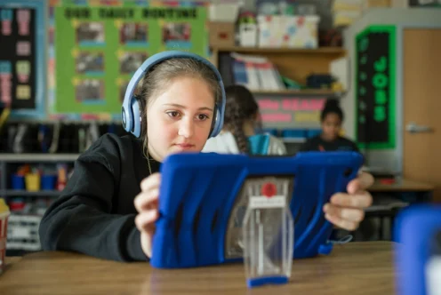 A student with headphones uses a tablet at a desk in a classroom, with shelves and a bulletin board visible in the background.