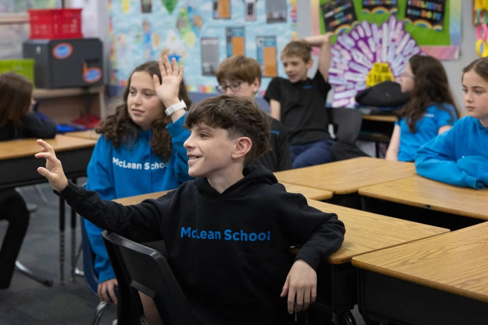 Students sit at desks in a classroom, with a boy in a "McLean School" hoodie raising his hand. Other students are also raising their hands or looking forward.