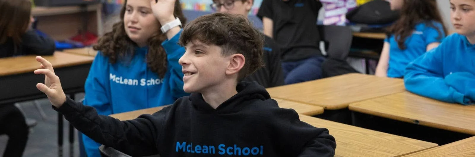 Students sit at desks in a classroom, with a boy in a "McLean School" hoodie raising his hand. Other students are also raising their hands or looking forward.