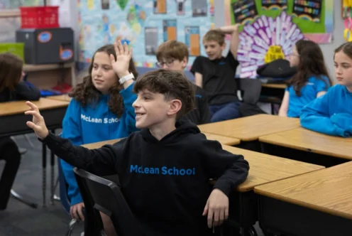 Students sit at desks in a classroom, with a boy in a "McLean School" hoodie raising his hand. Other students are also raising their hands or looking forward.