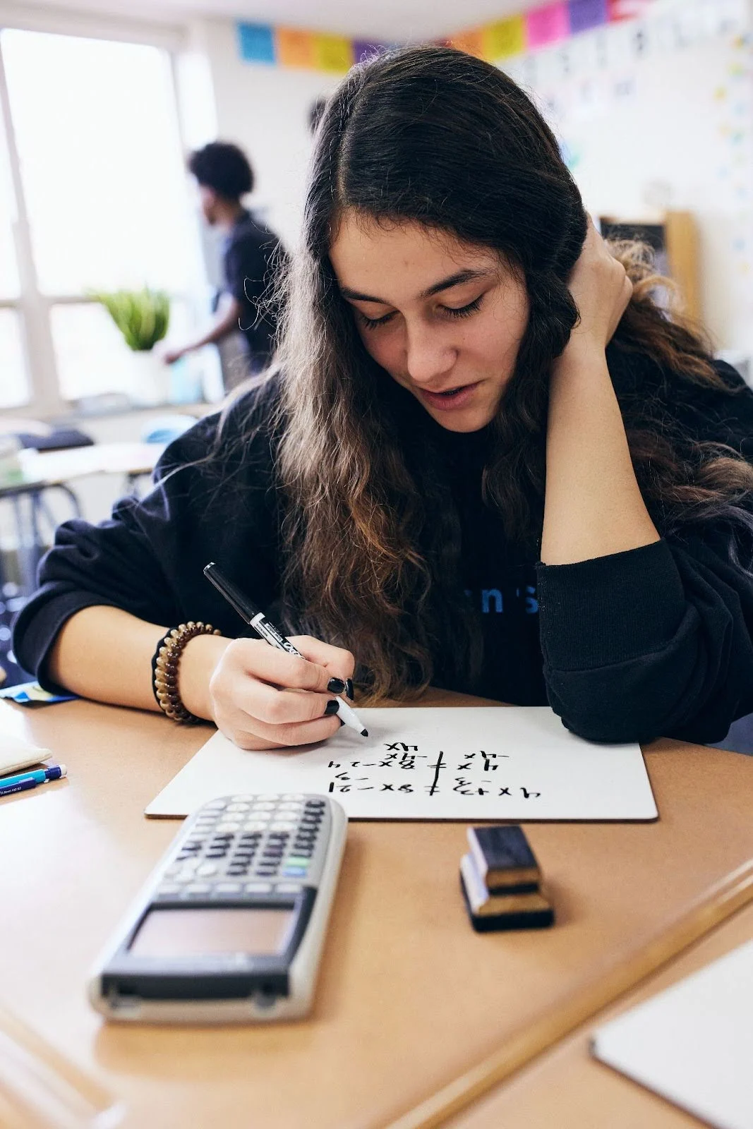 A student with long hair writes on a whiteboard at a desk with a calculator and eraser, in a classroom setting.