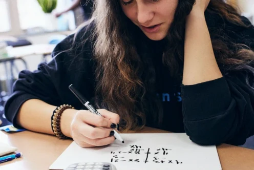 A student with long hair writes on a whiteboard at a desk with a calculator and eraser, in a classroom setting.