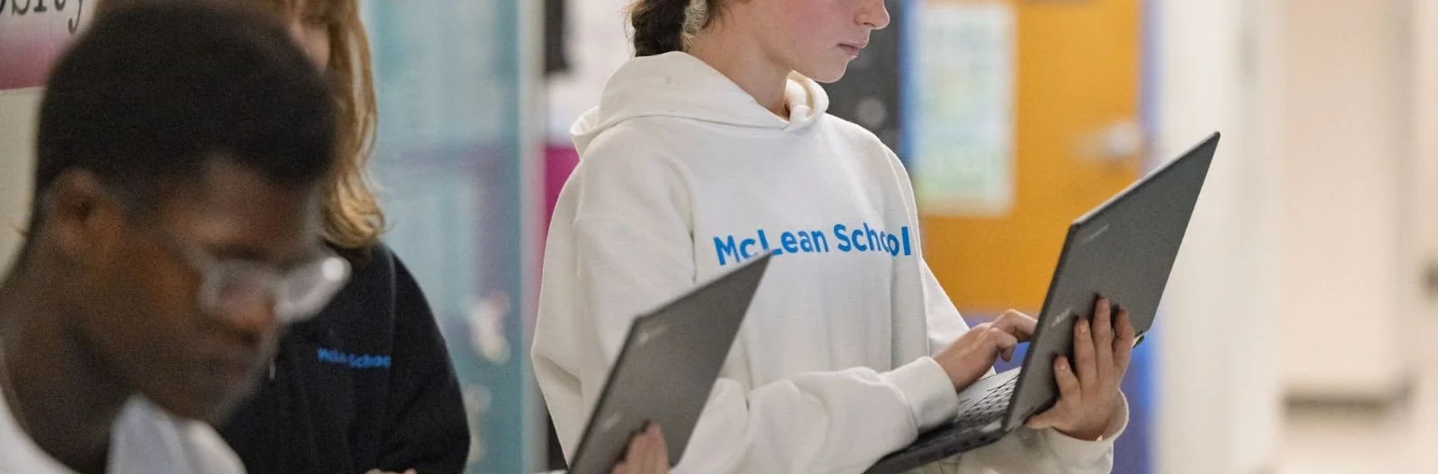 Three students stand in a school hallway holding and using laptops. The student in front is wearing a white McLean School hoodie.